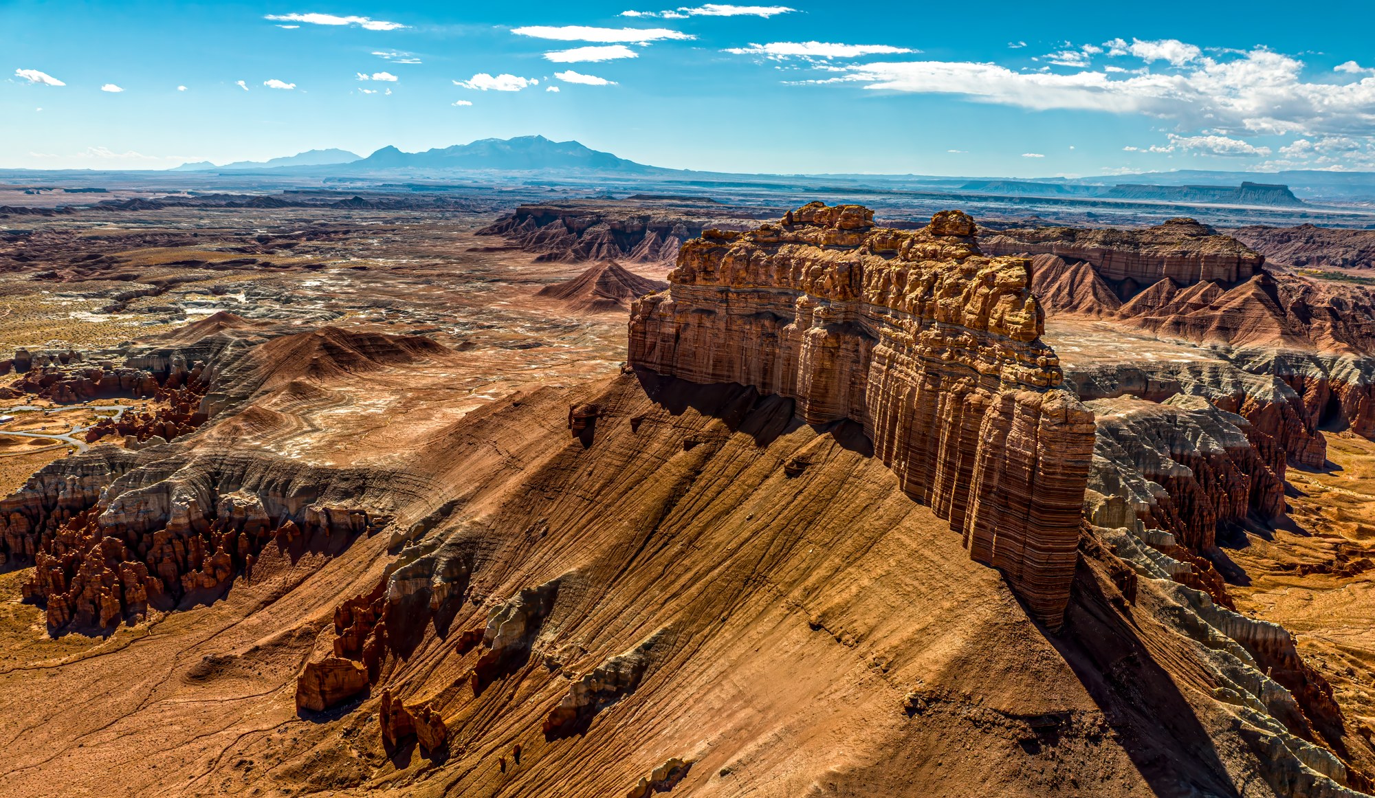 Aerial view of a banded sandstone butte rising above painted desert badlands, distant blue mountains on the horizon under a bright sky