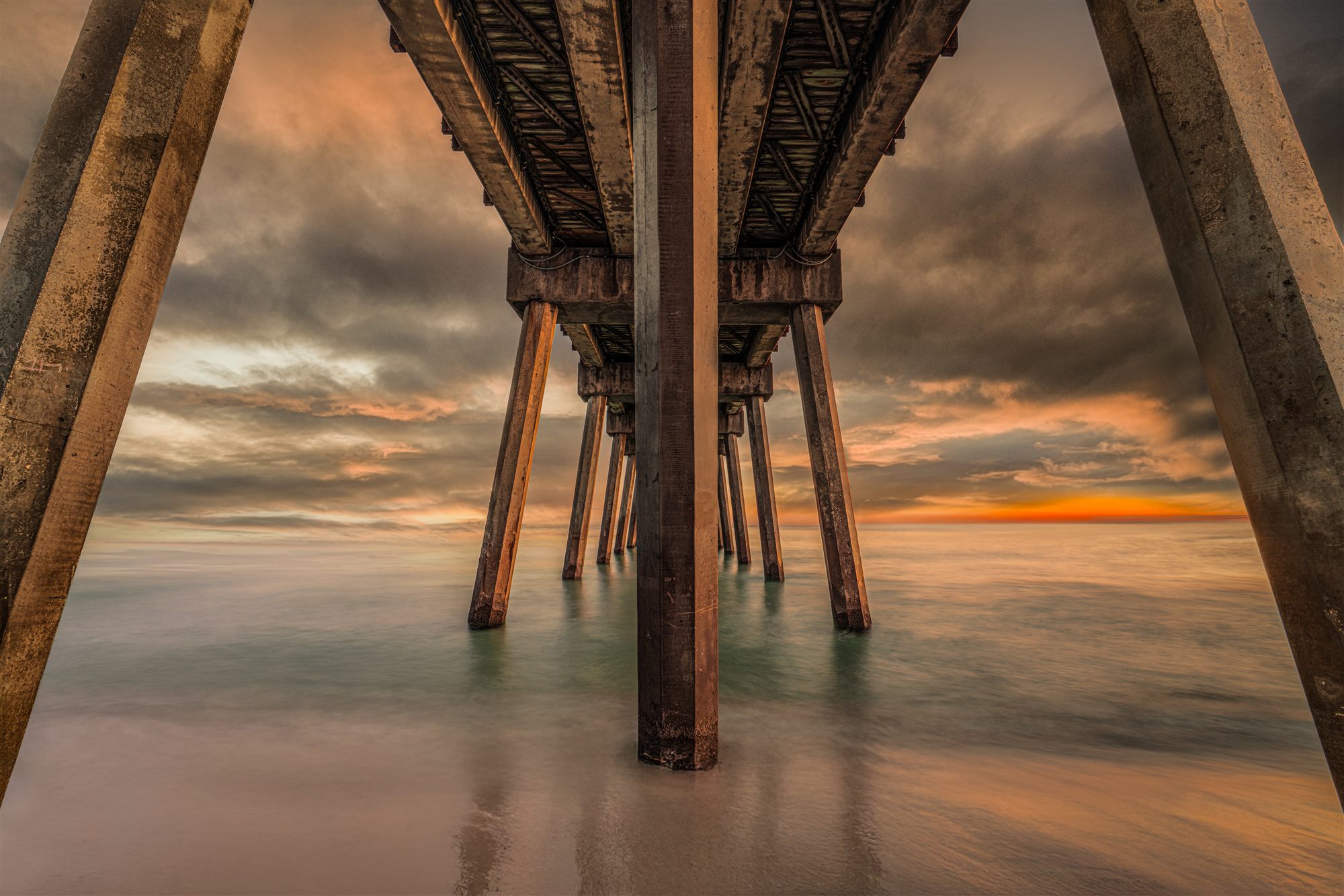 Long-exposure view beneath a pier at sunset, pilings receding to a burning horizon