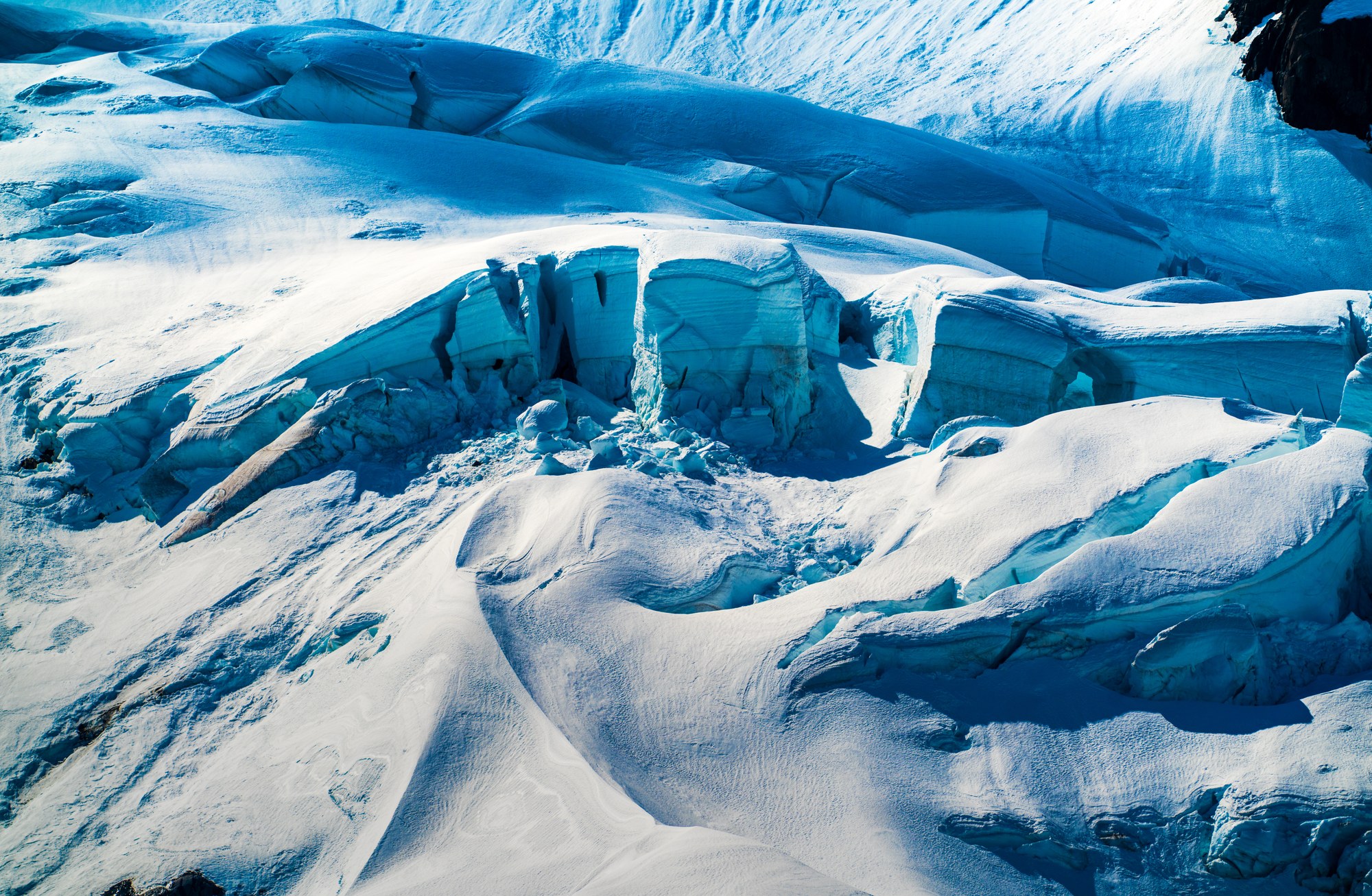 Glacier surface with broken blue seracs and crevasses emerging from wind-sculpted snow, dark rock at the edges