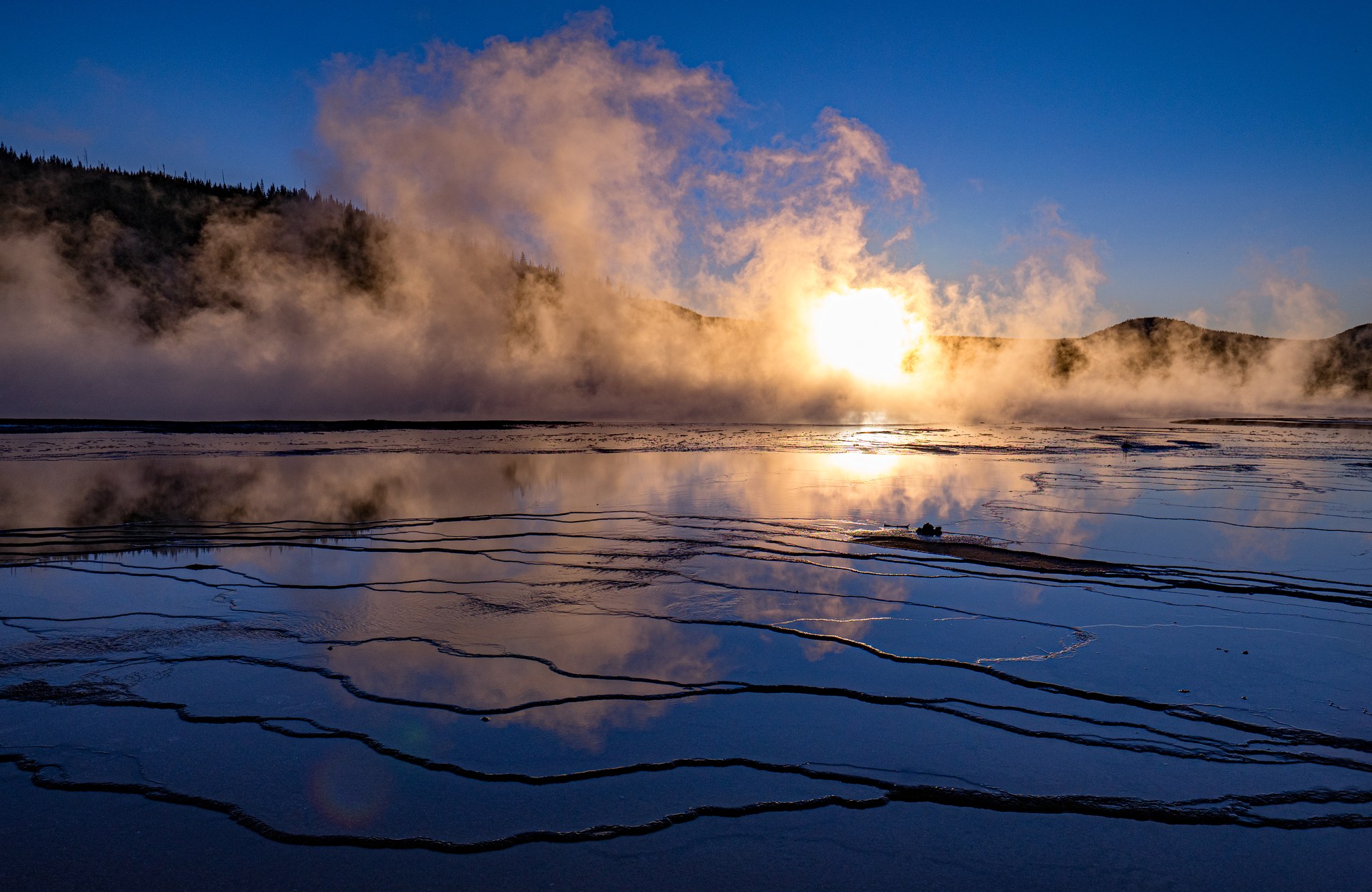 Backlit steam rising from a hot spring, the low sun glowing through the vapor, terraced mineral pools reflecting the blue sky in the foreground