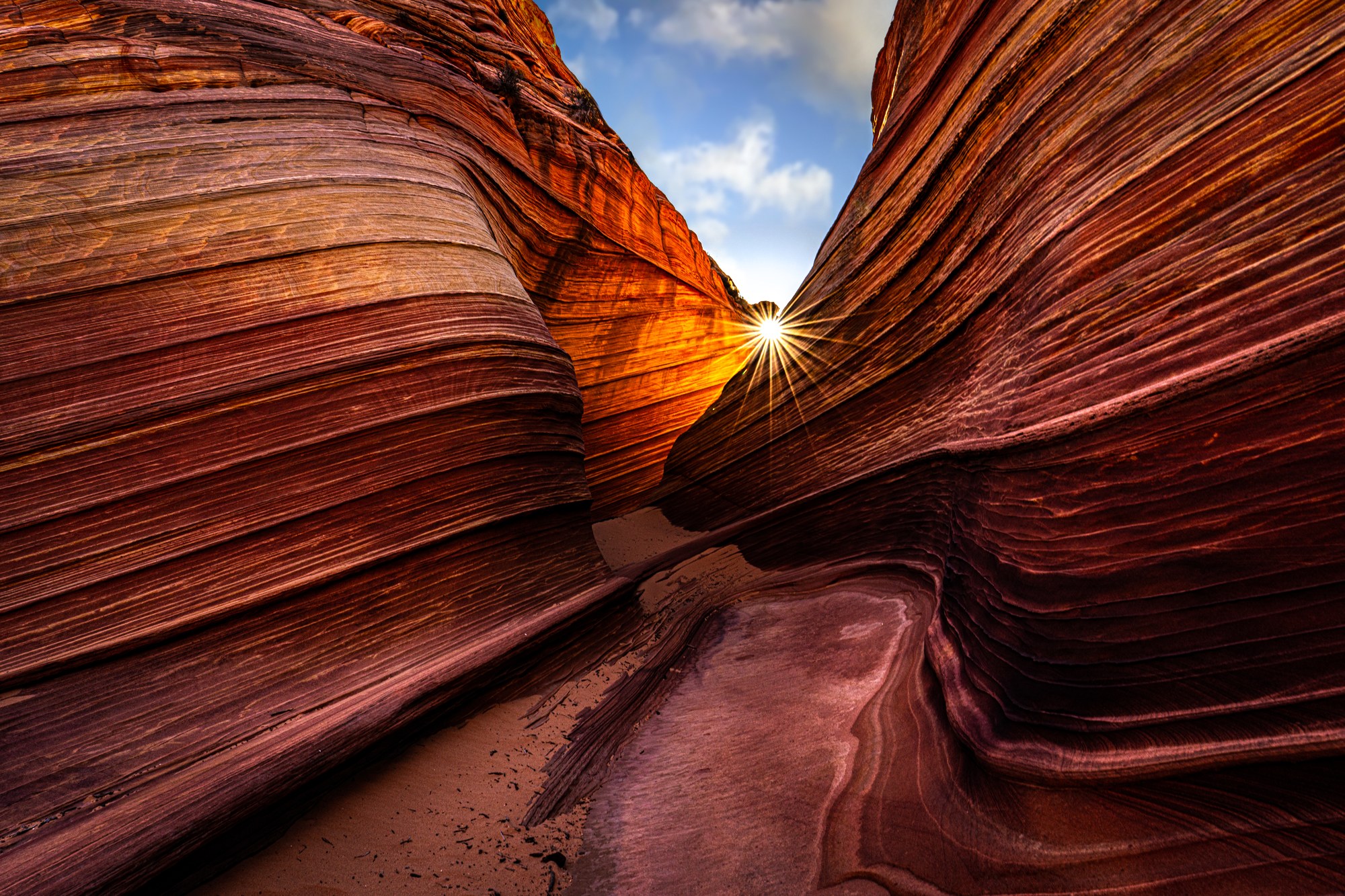 Striped swirling sandstone walls of The Wave at Coyote Buttes, a sunstar bursting through the notch between them under a blue sky