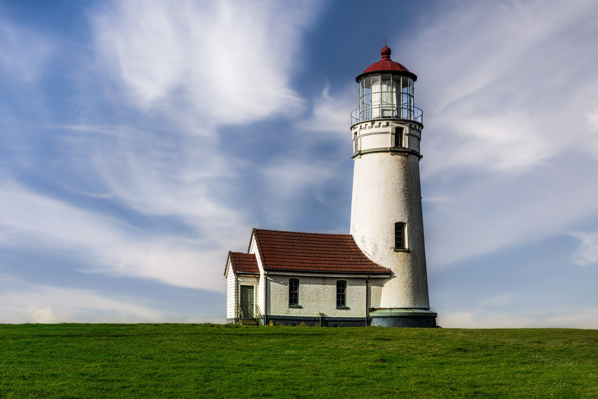 White lighthouse with a red lantern roof and attached keeper's building standing on a green lawn under wispy white clouds