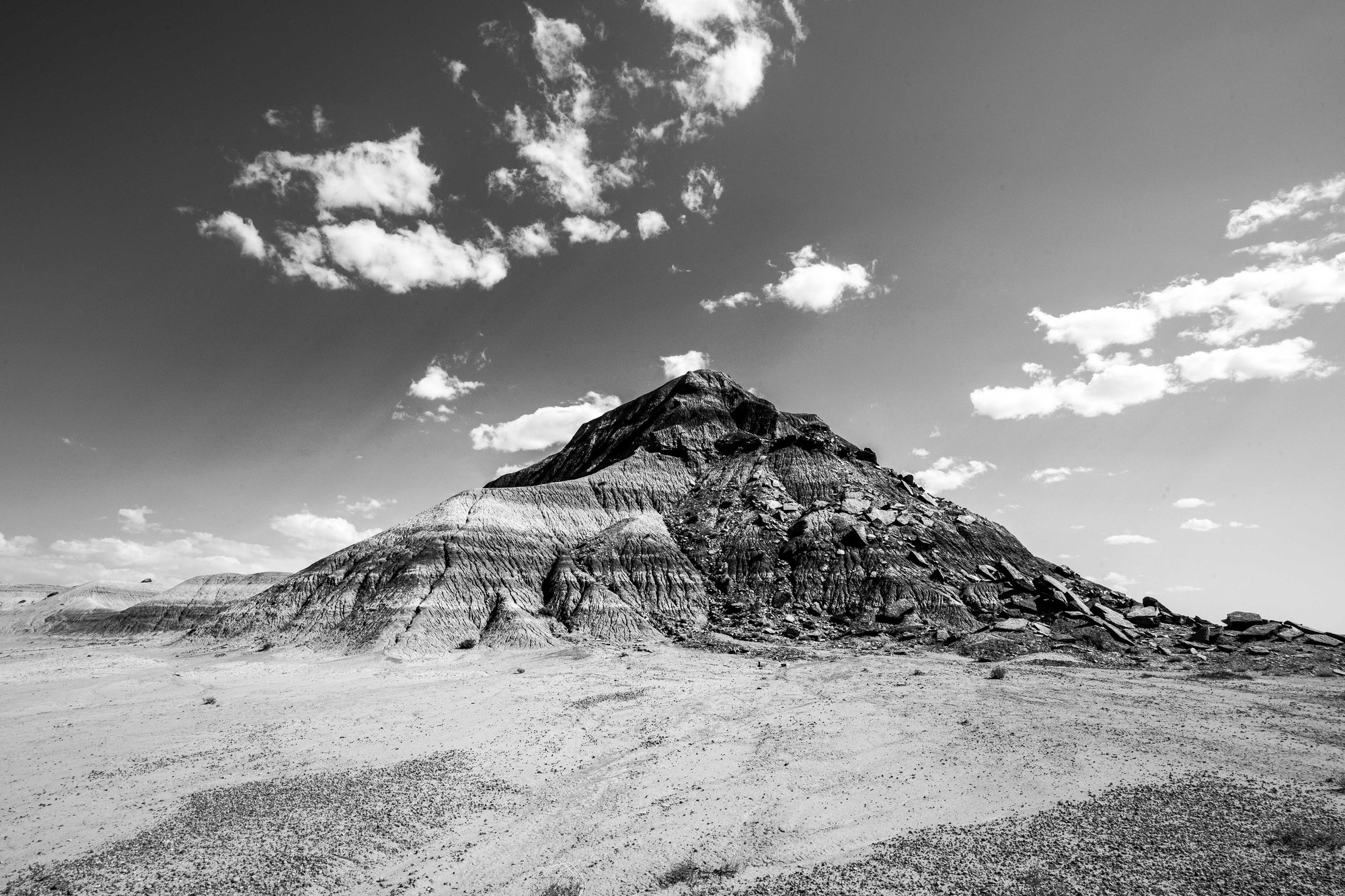 Black-and-white banded badlands mound with broken rock on its slopes, barren desert floor in front and small clouds in a dark sky