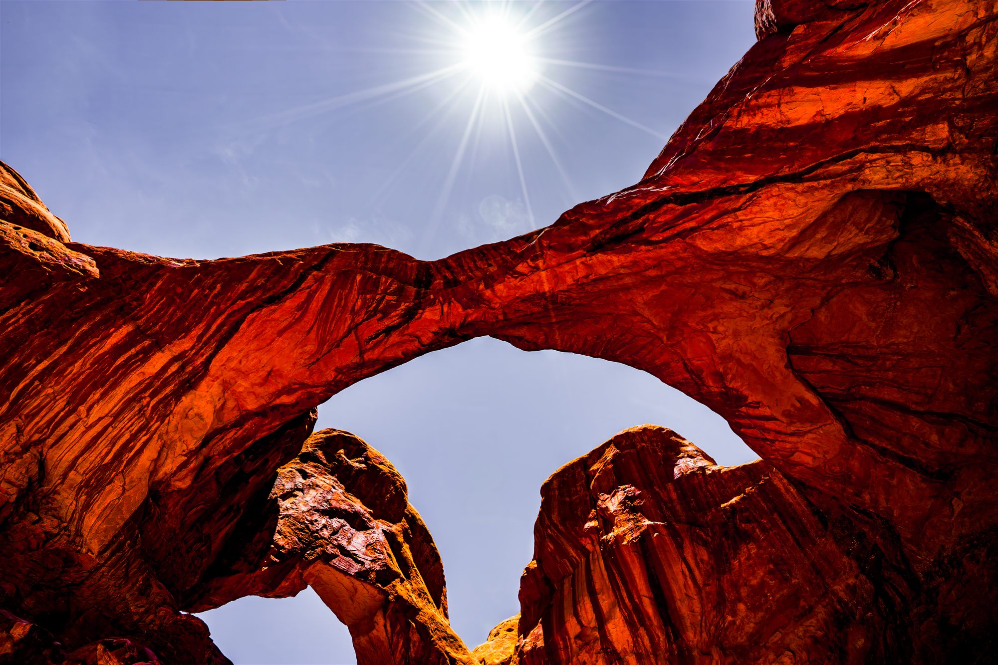 Double sandstone arch glowing red against a blue sky, the midday sun flaring above it