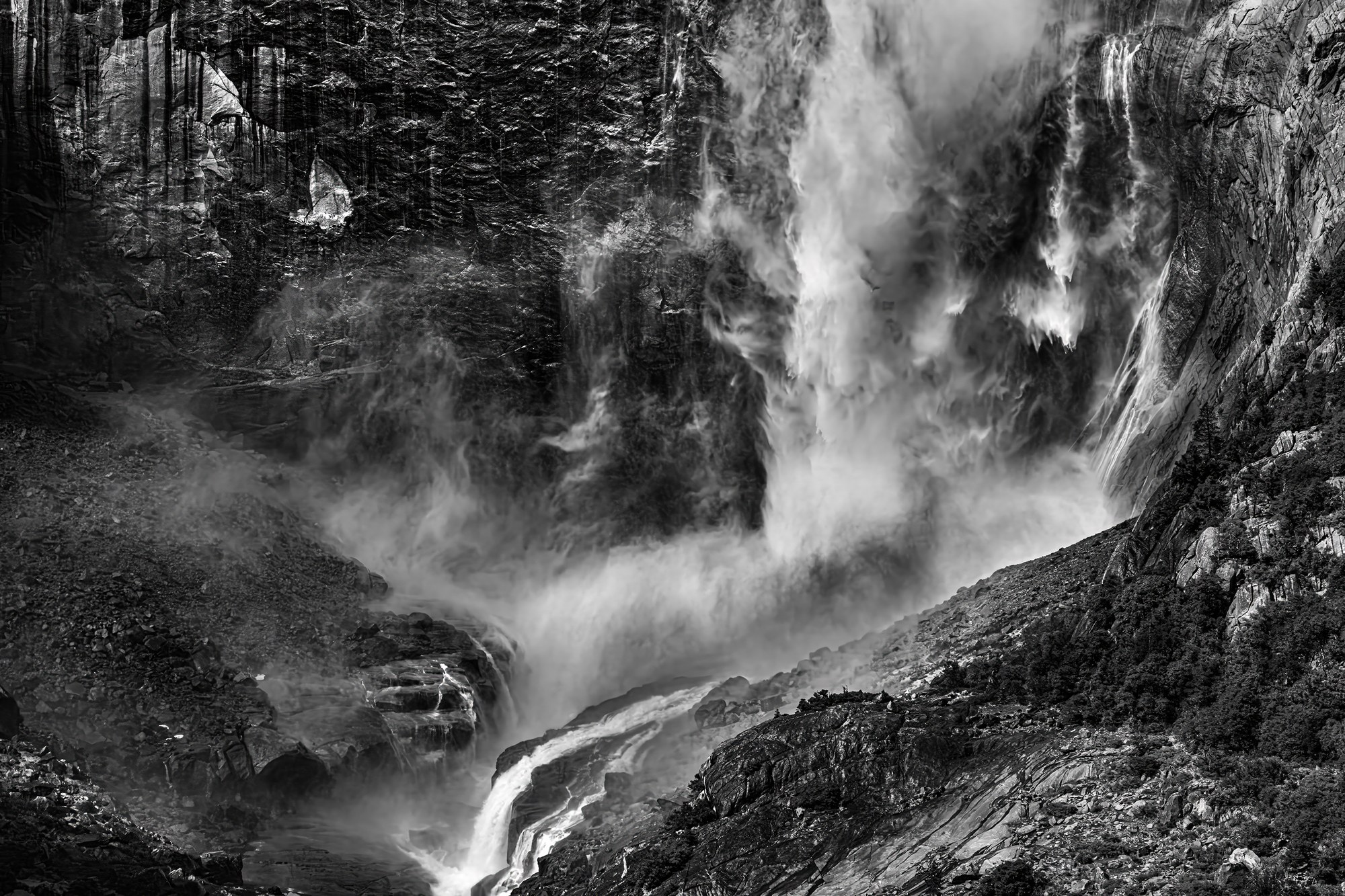 Black-and-white waterfall crashing down a dark granite cliff into a boulder-strewn gorge, mist billowing upward