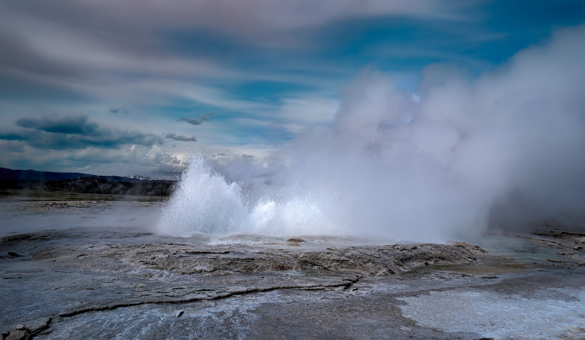 Geyser erupting from a white mineral-crusted basin, a tall column of water and steam against a blue sky with streaked clouds