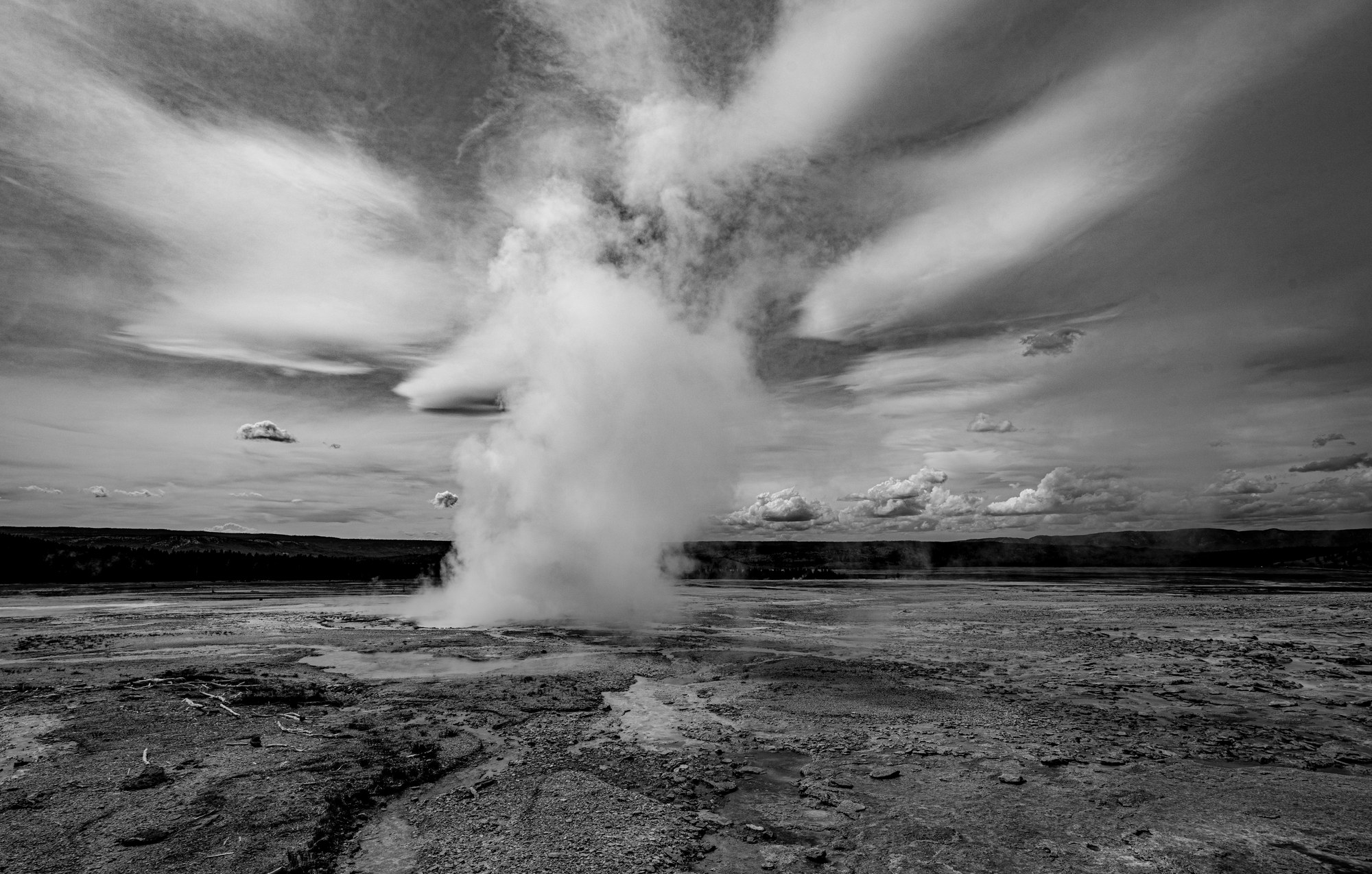 Black-and-white geyser basin with a tall column of steam rising from a barren sinter flat and merging into streaked clouds overhead