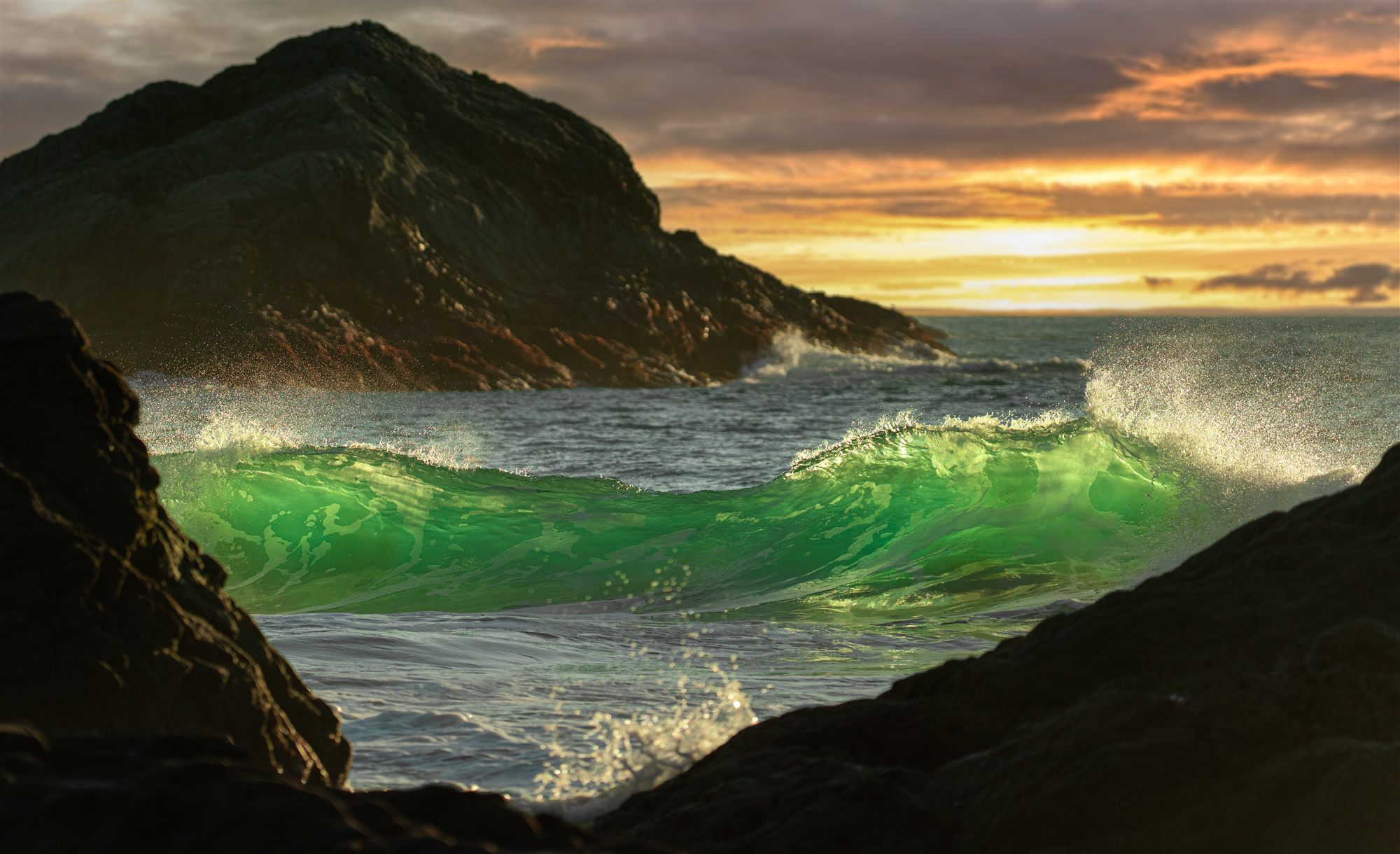 Emerald-green wave lit from within by a golden sunset, framed by dark coastal rocks