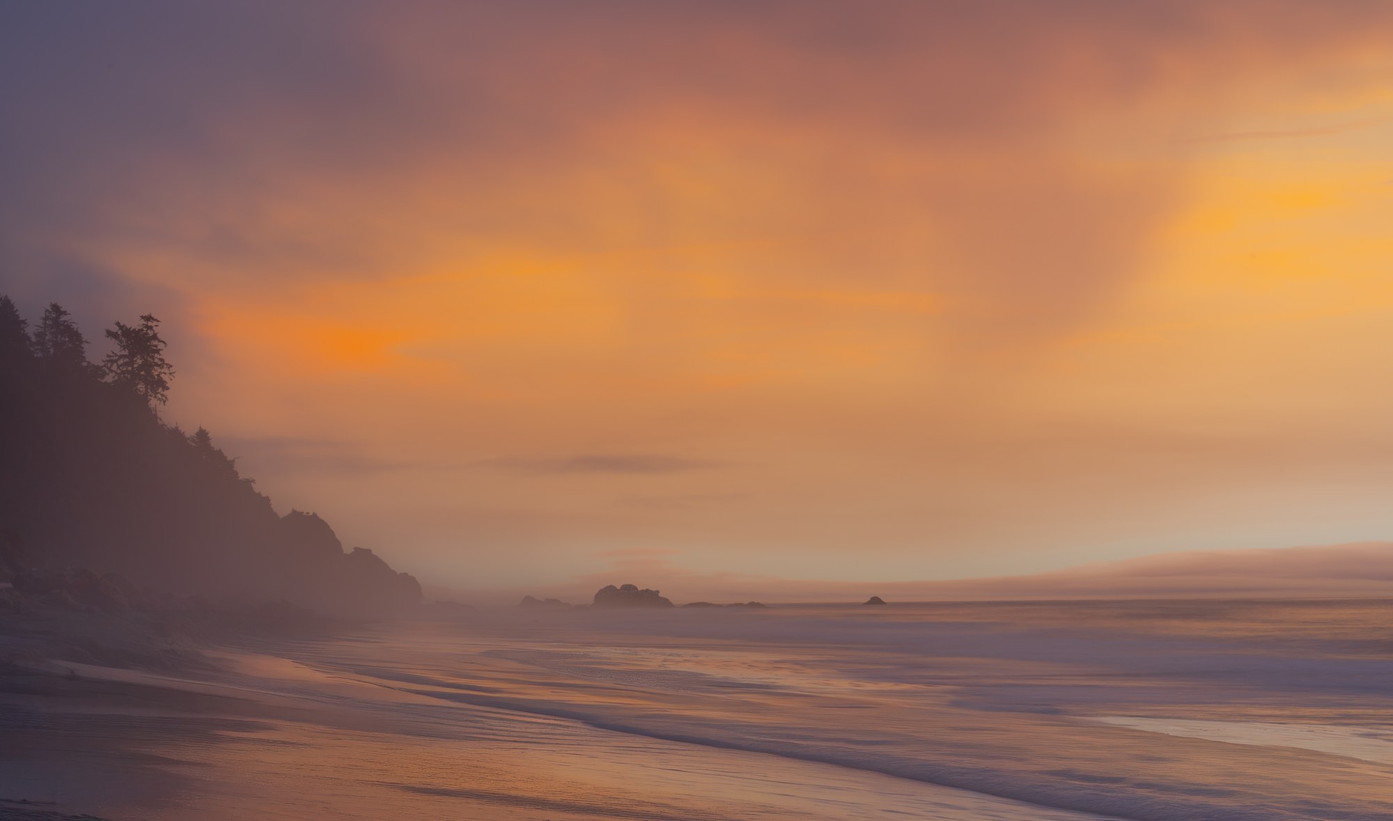 Misty beach at sunset in soft peach and lavender light, a dark forested headland at left and sea stacks fading into haze along the tideline