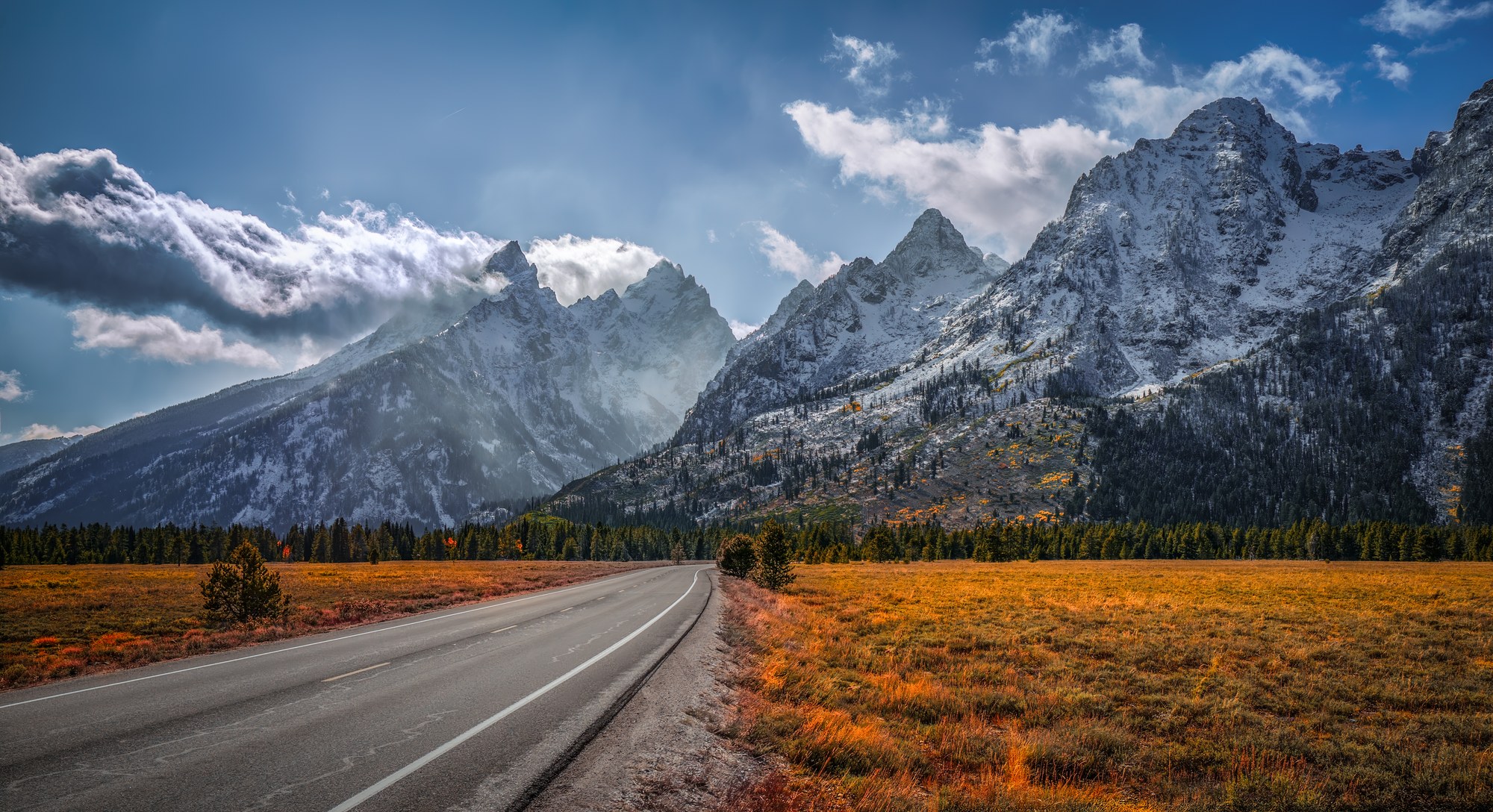 Empty two-lane road curving toward snow-dusted Teton peaks under dramatic clouds, autumn-gold meadow grass on either side