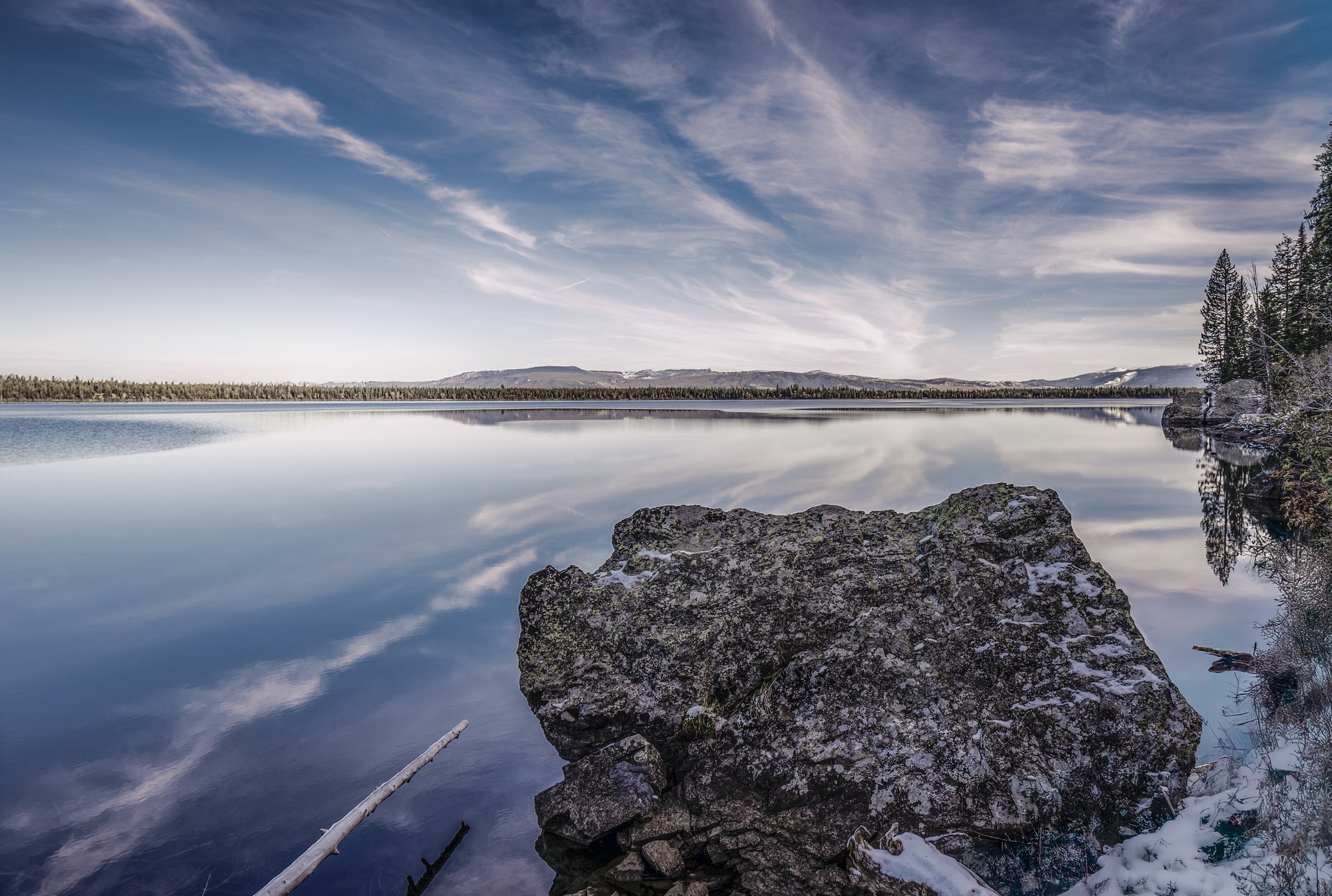 Calm lake mirroring wispy clouds, a large lichen-covered boulder dusted with snow on the shore, forested far shoreline and distant hills