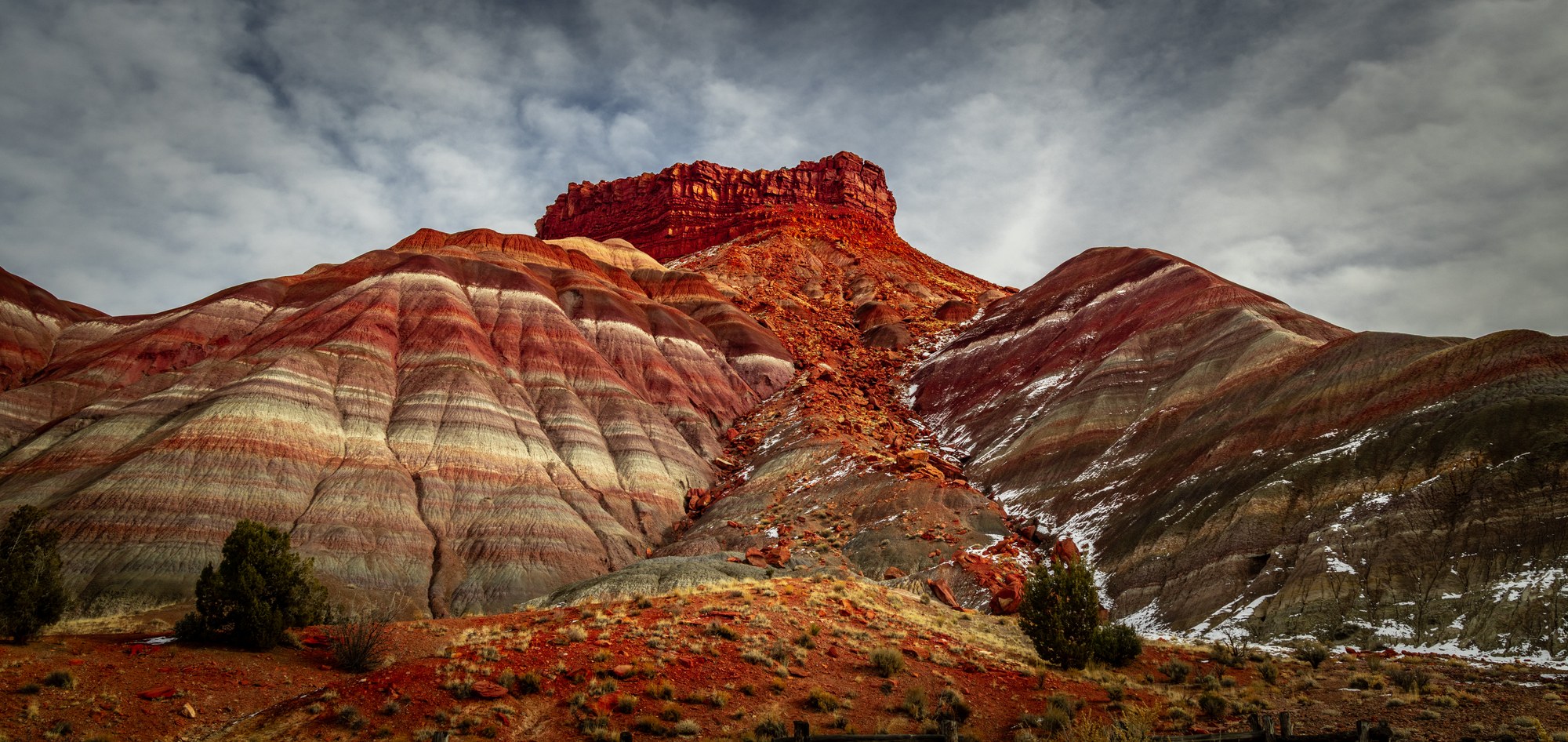 Striped red, pink and grey banded badlands hills capped by a dark red rock butte under a cloudy sky, patches of snow in the gullies