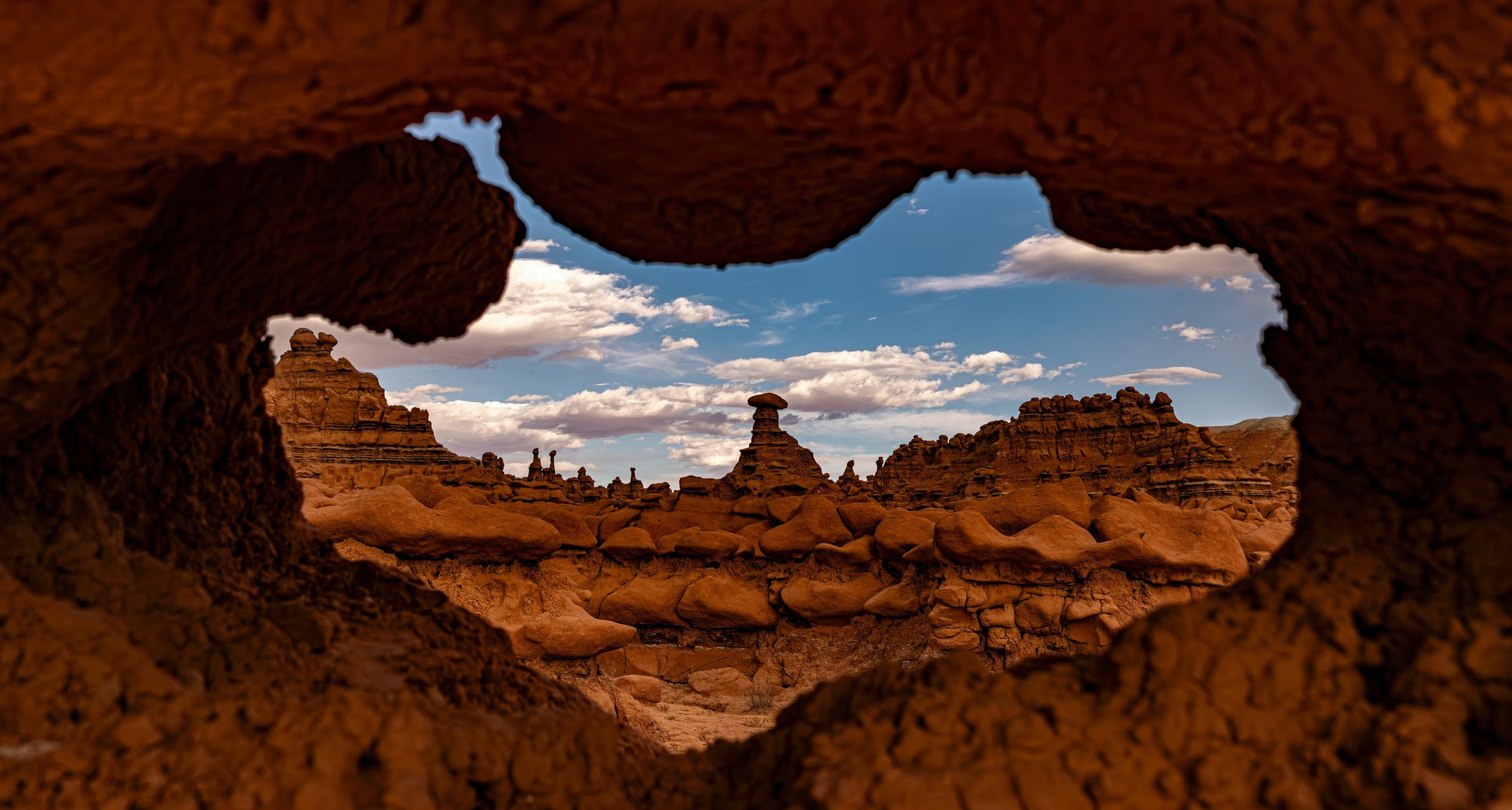 View through an eroded hole in knobby red rock toward mushroom-shaped hoodoos of Goblin Valley, Utah, under a blue sky with clouds