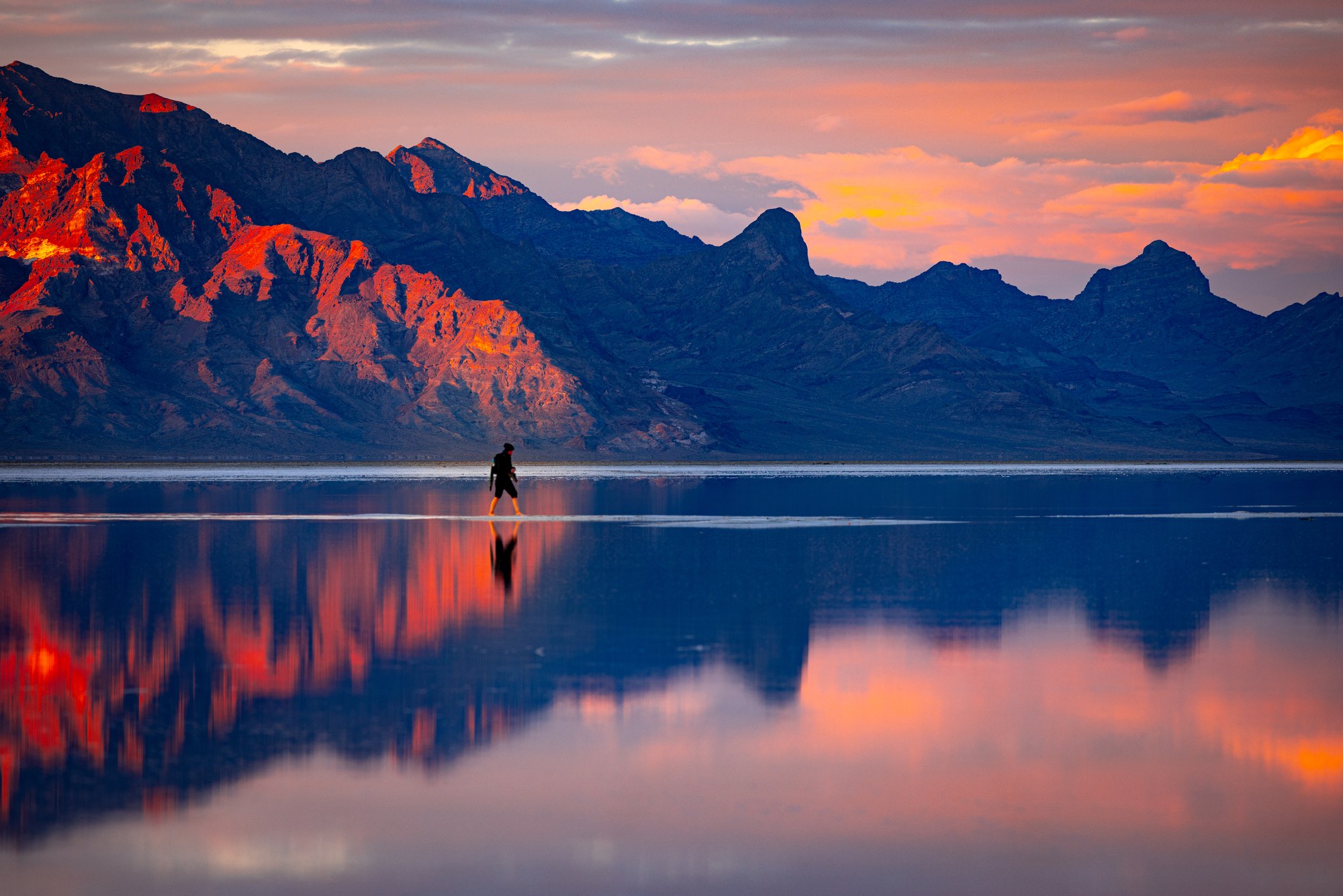 Lone person walking across flooded salt flats at sunset, red alpenglow on the mountains behind, clouds mirrored in the shallow water