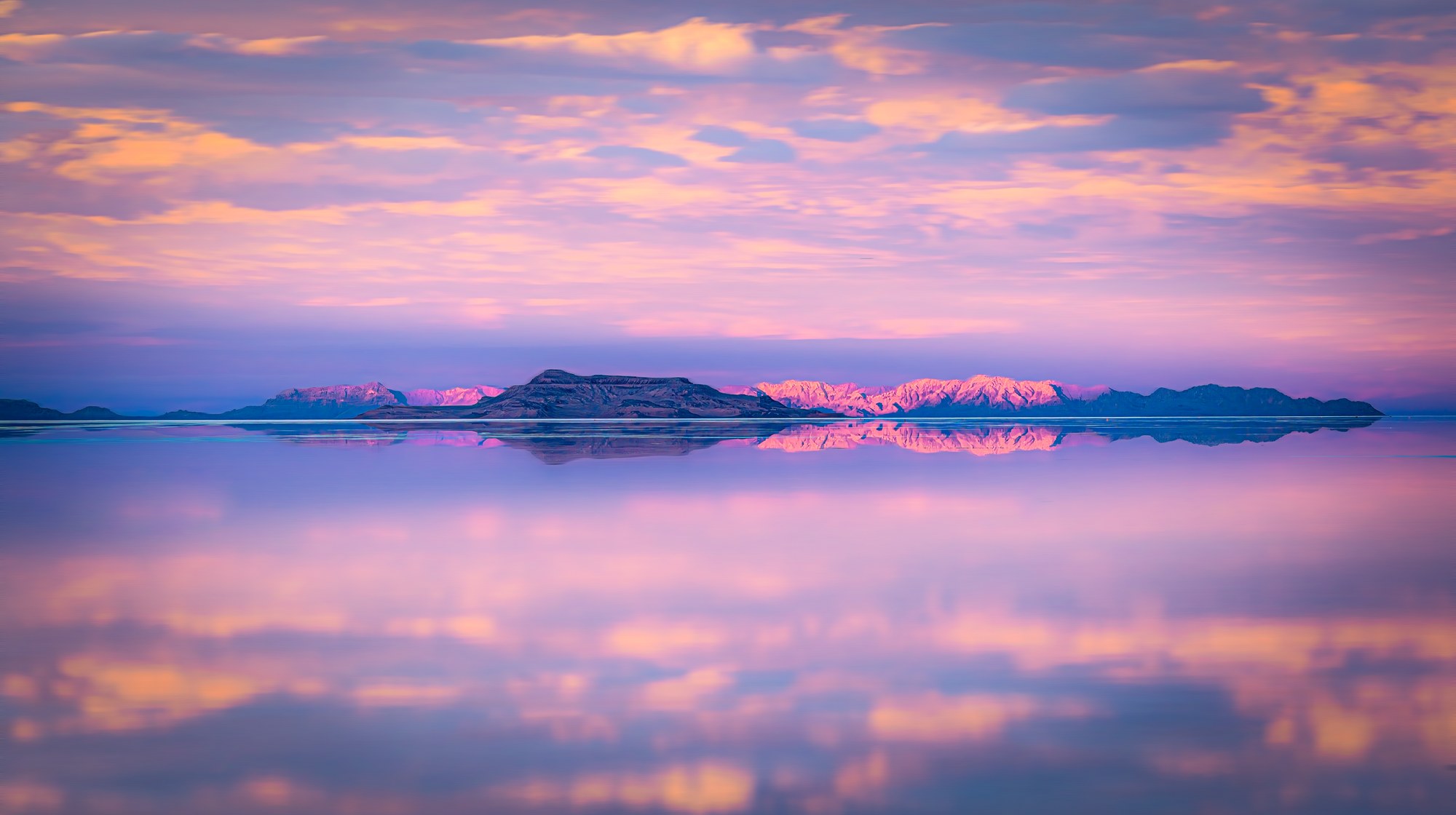 Flooded salt flats mirroring a pink and gold sunset sky, distant snow-capped mountains reflected along the horizon