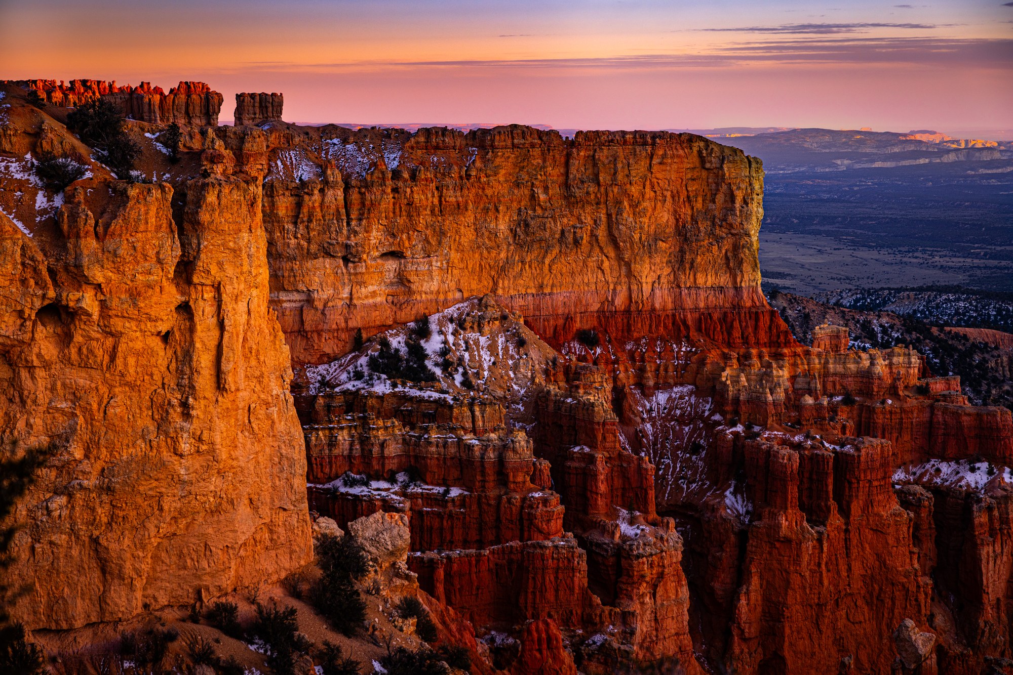 Glowing orange cliffs and hoodoos of Bryce Canyon at sunset, patches of snow in the shadows, a distant valley fading to blue under a pink sky